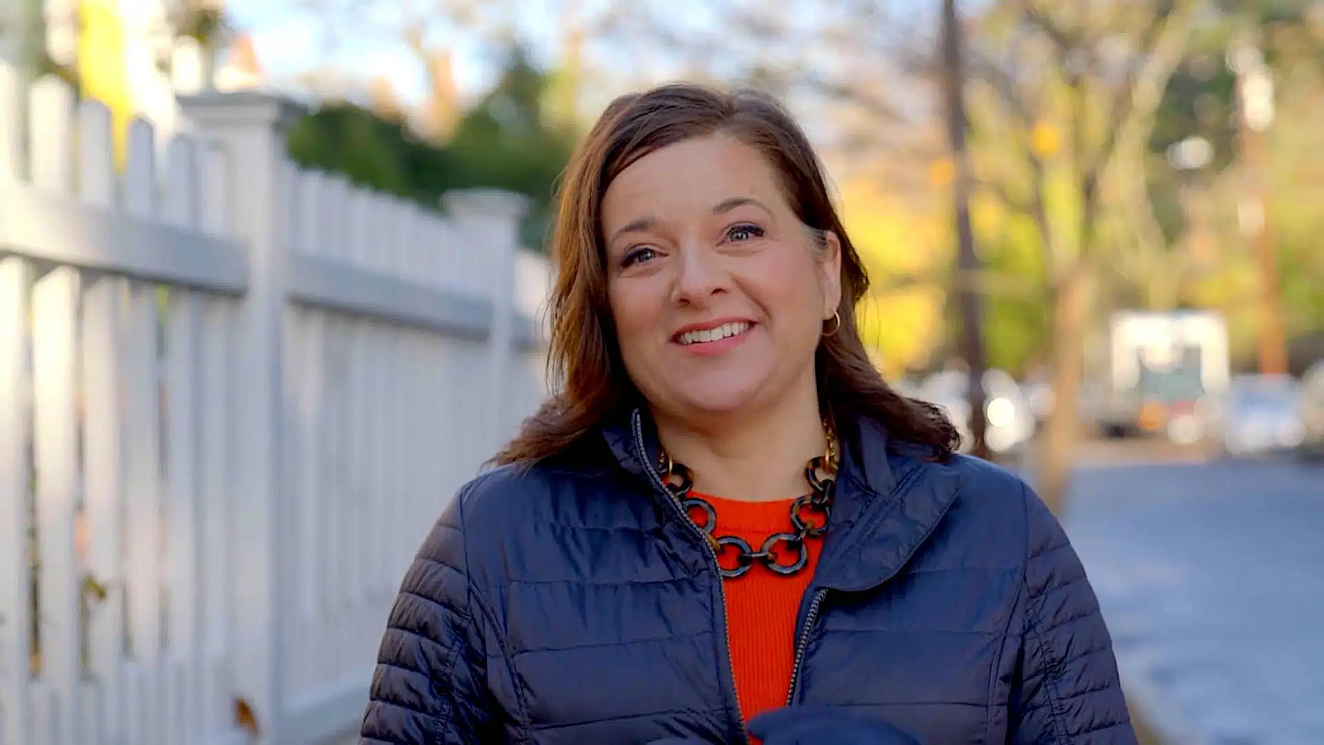 Person wearing a dark jacket and orange shirt, standing outdoors near a white fence on a sunny day, smiling at the camera.