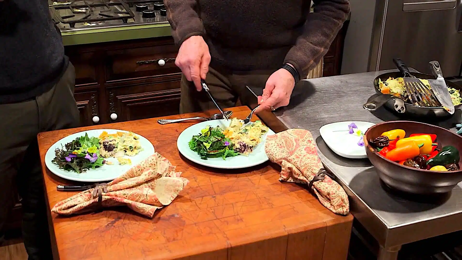 A person serves food onto a plate in a kitchen. In front of them are two plates with food, two patterned napkins, and a bowl of colorful vegetables. A stovetop is in the background.