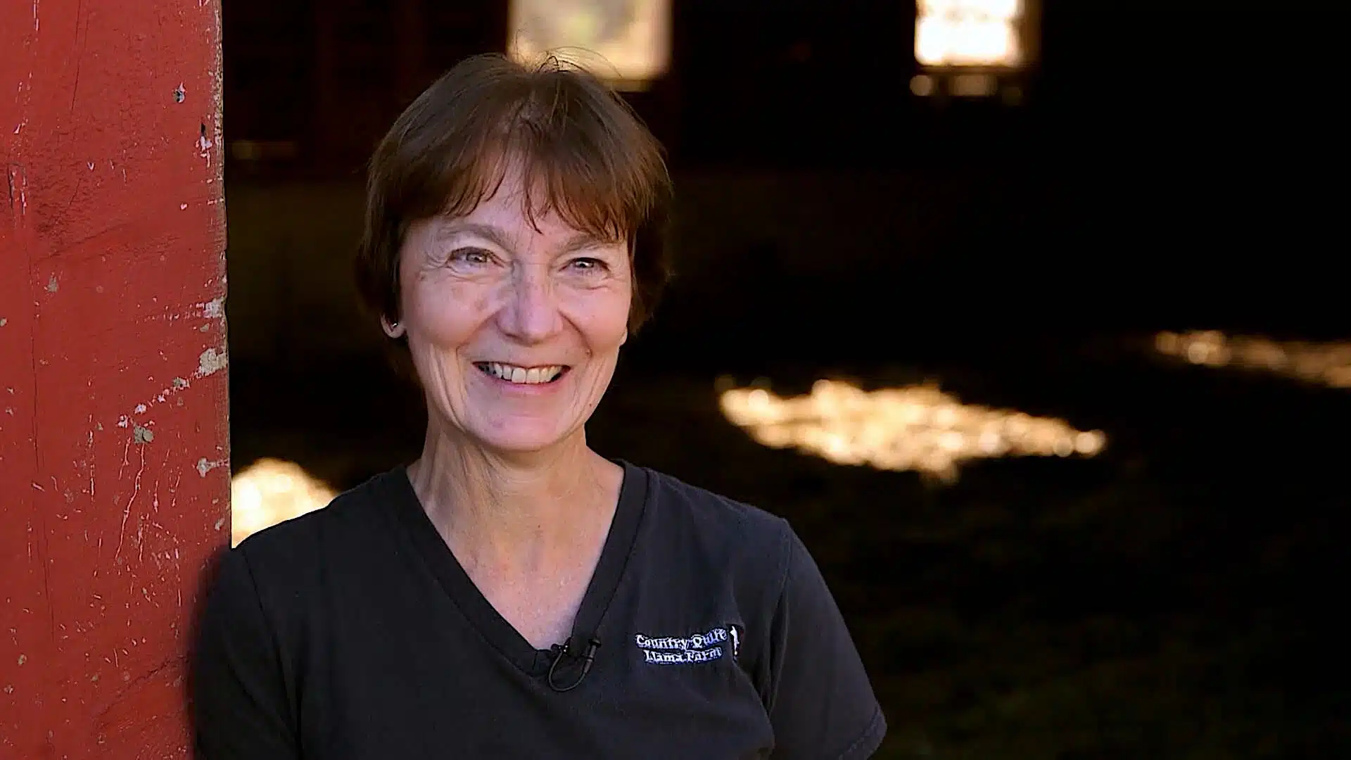 A woman with short brown hair in a black T-shirt stands next to a red wall, smiling at the camera. The background is dimly lit.