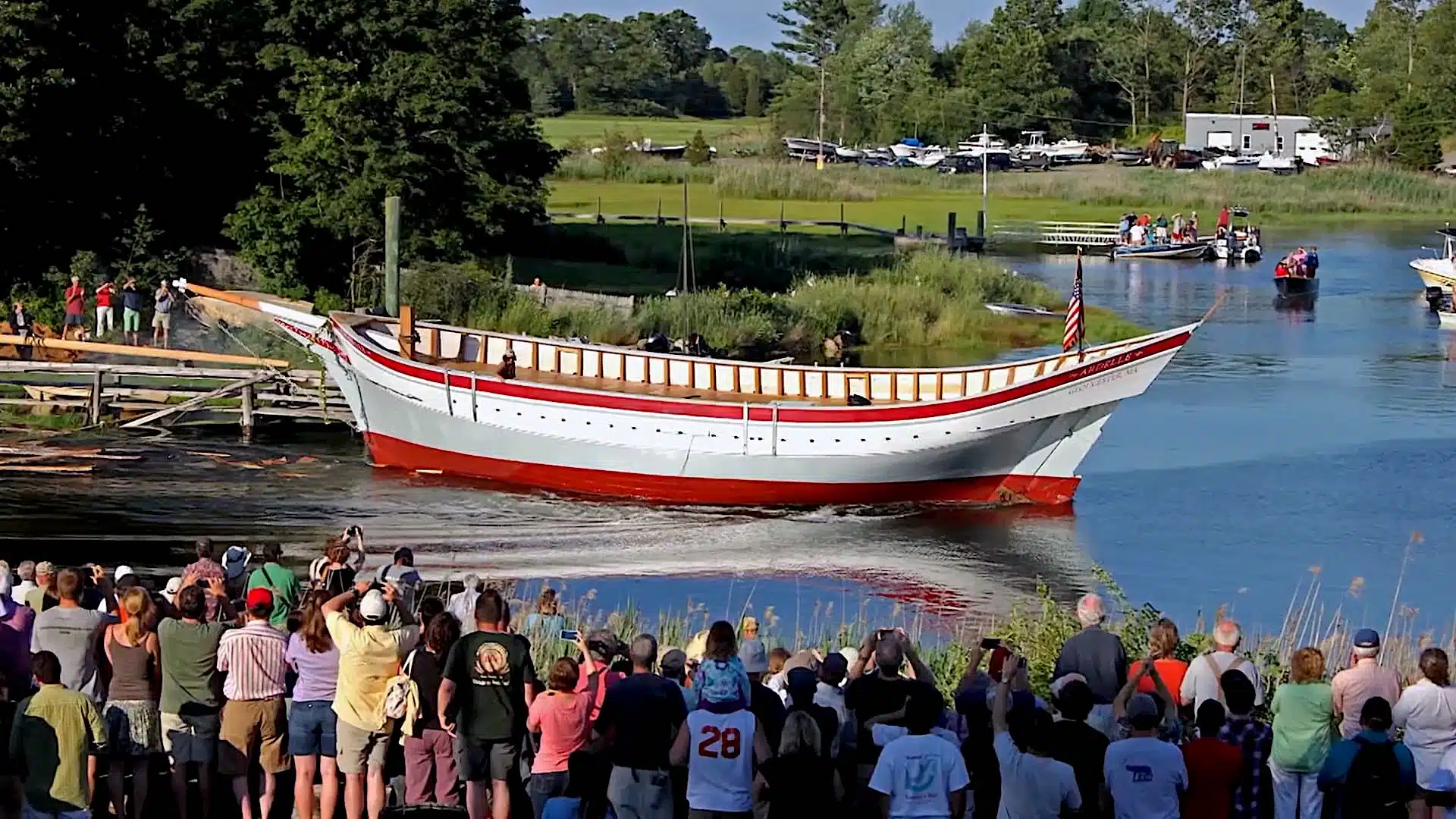 A crowd watches as a newly constructed boat is launched into the water in a grassy, wooded area with various onlookers and parked cars in the distance.