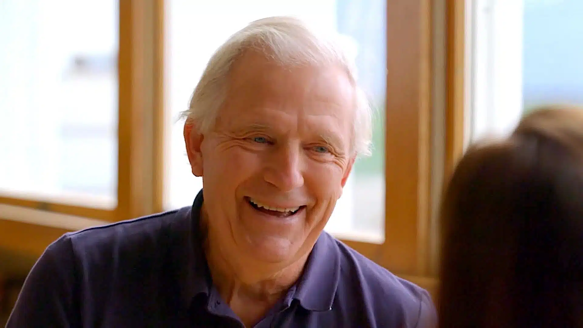 A smiling elderly man with white hair and a navy blue polo shirt sits near a window, conversing with a person whose back is visible in the foreground.