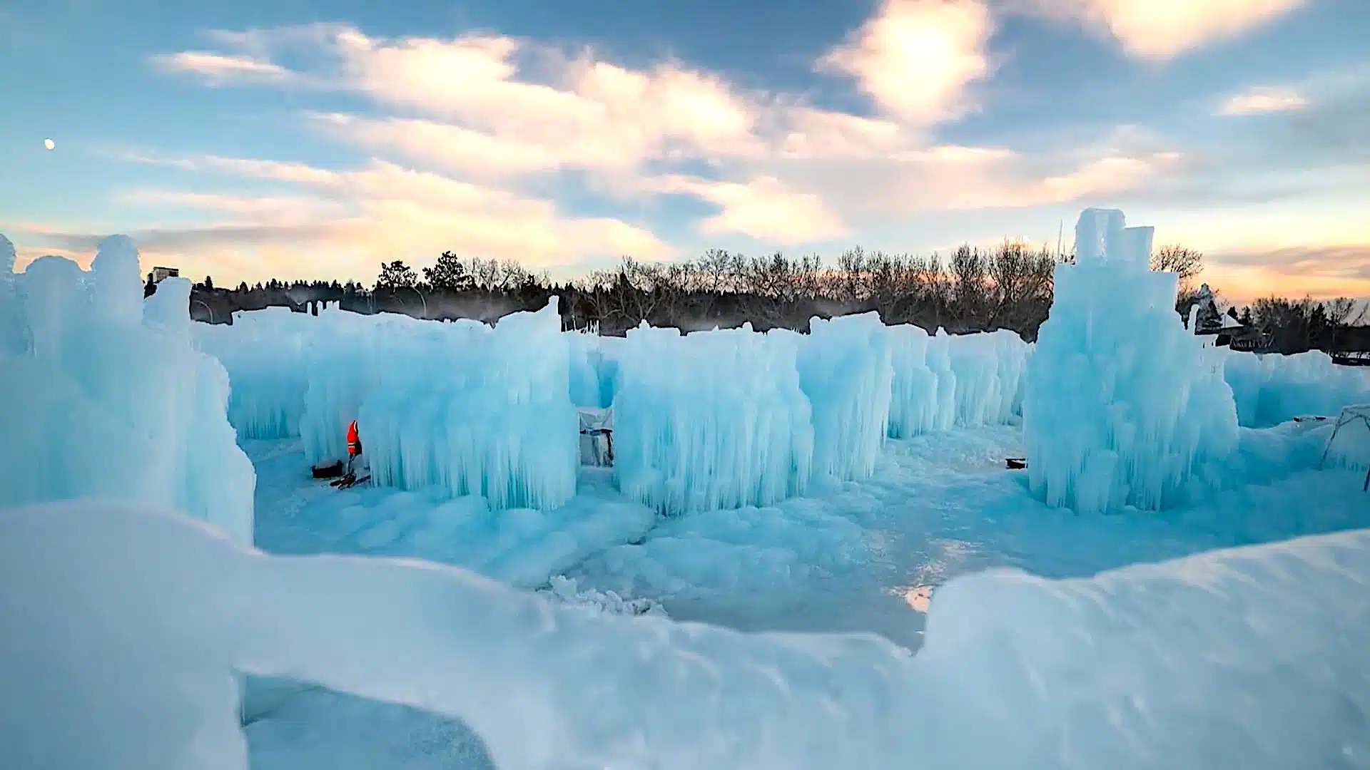 A winter landscape featuring large ice formations under a blue sky with clouds. A person in an orange jacket stands amidst the ice structures. Trees are visible in the background.