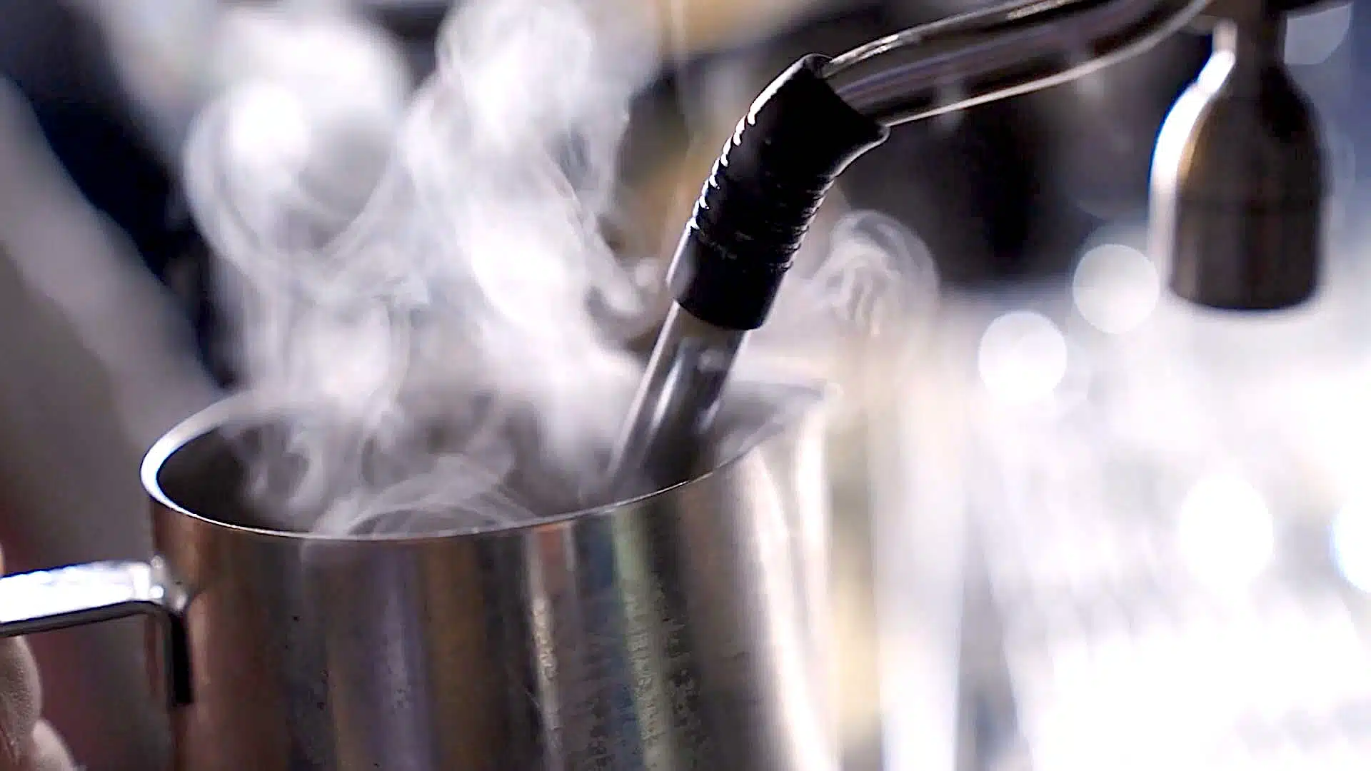 A steam wand froths milk in a stainless steel pitcher. Steam is visible rising from the milk.
