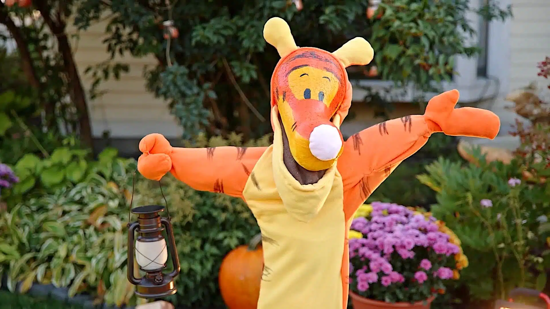 gourd in a Tigger costume holding a lantern stands in a colorful garden with various plants and flowers, including a pumpkin in the background.