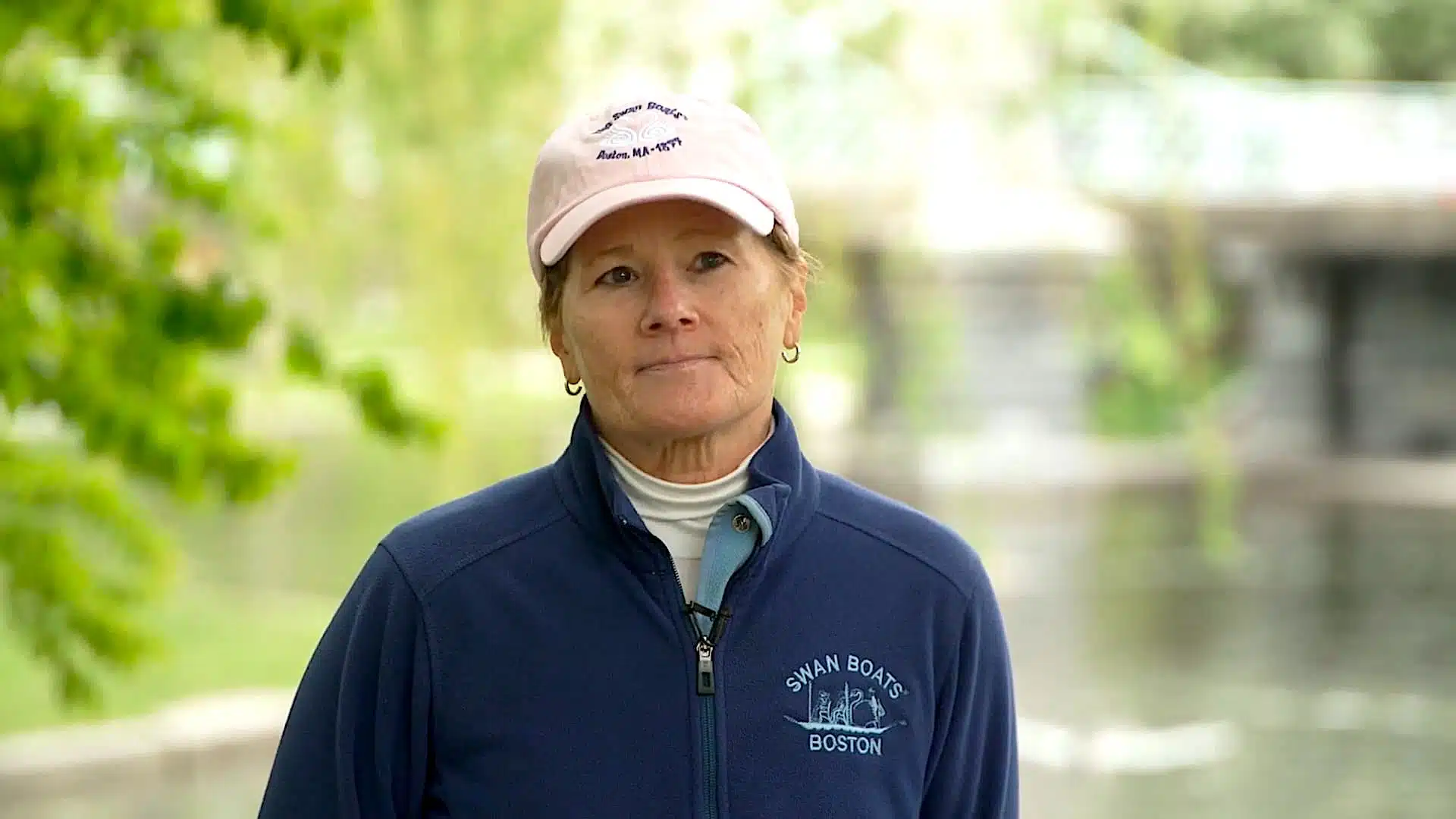 Person wearing a pink cap and a blue jacket with "Swan Boats Boston" standing outdoors near a body of water.