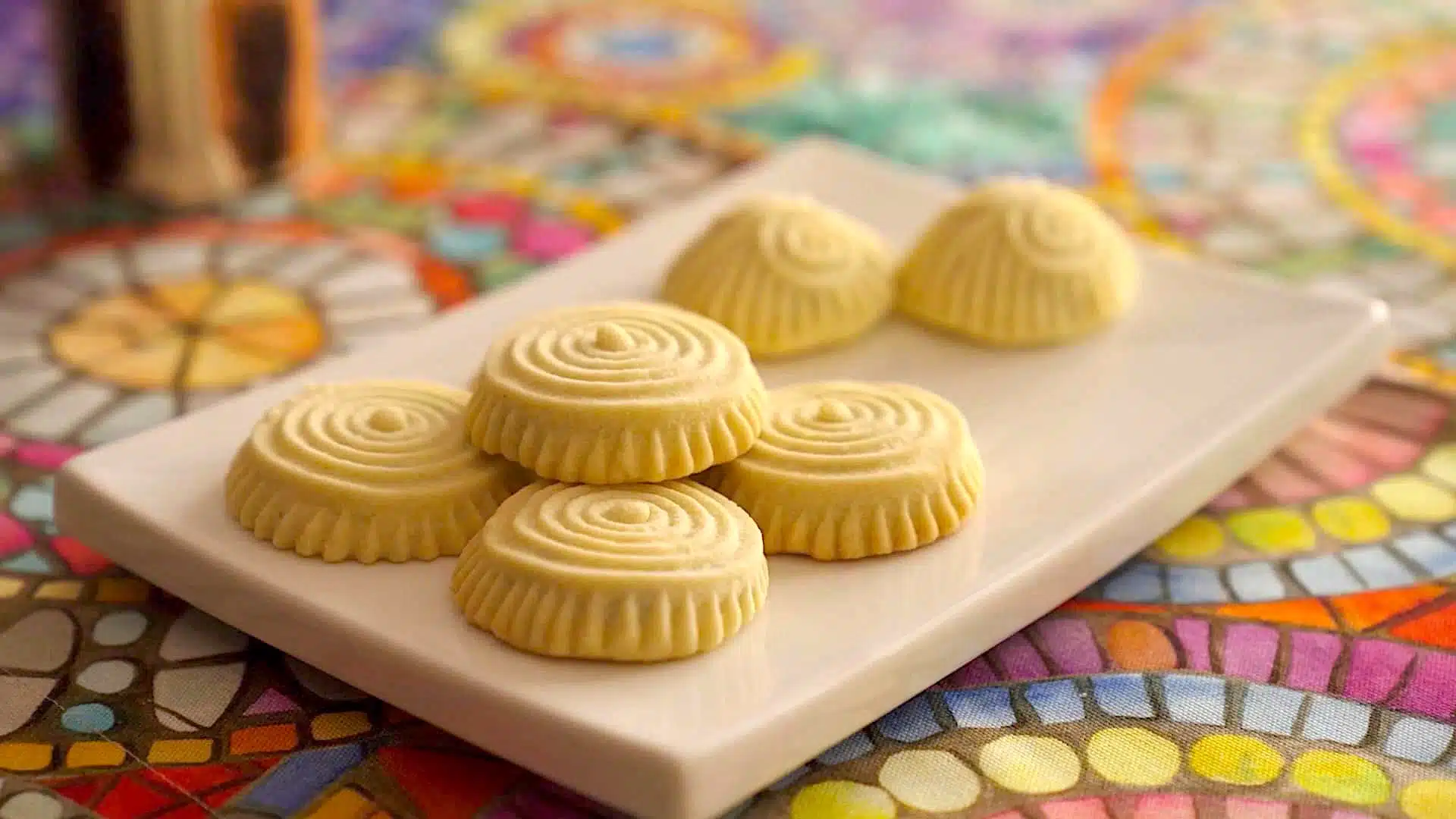 A white rectangular plate with six round cookies patterned with swirls on a colorful, mosaic-patterned tablecloth.