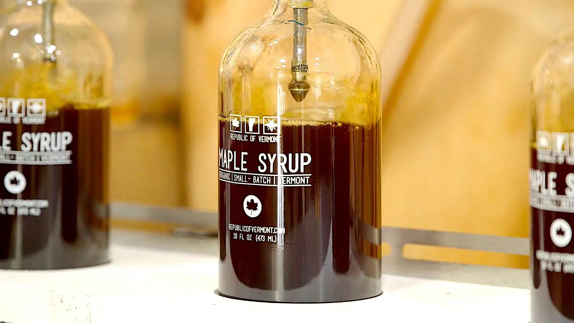 Clear glass bottles labeled "Maple Syrup" filled with dark liquid, placed on a white counter, with a metal dispenser inserted into the top of one bottle.
