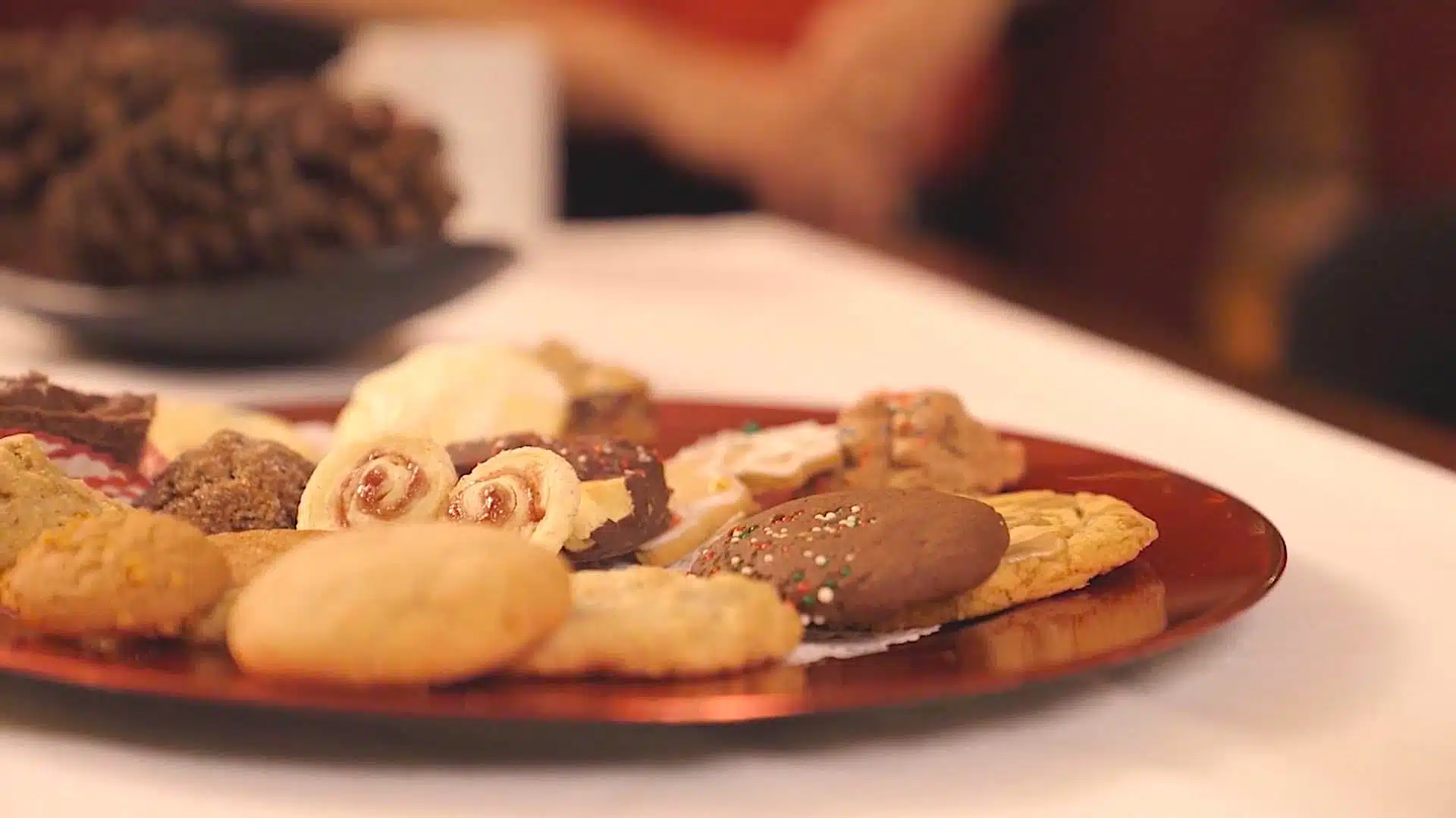 A variety of cookies and pastries are arranged on a red plate, placed on a white tablecloth. A blurred background shows more items and part of a person.