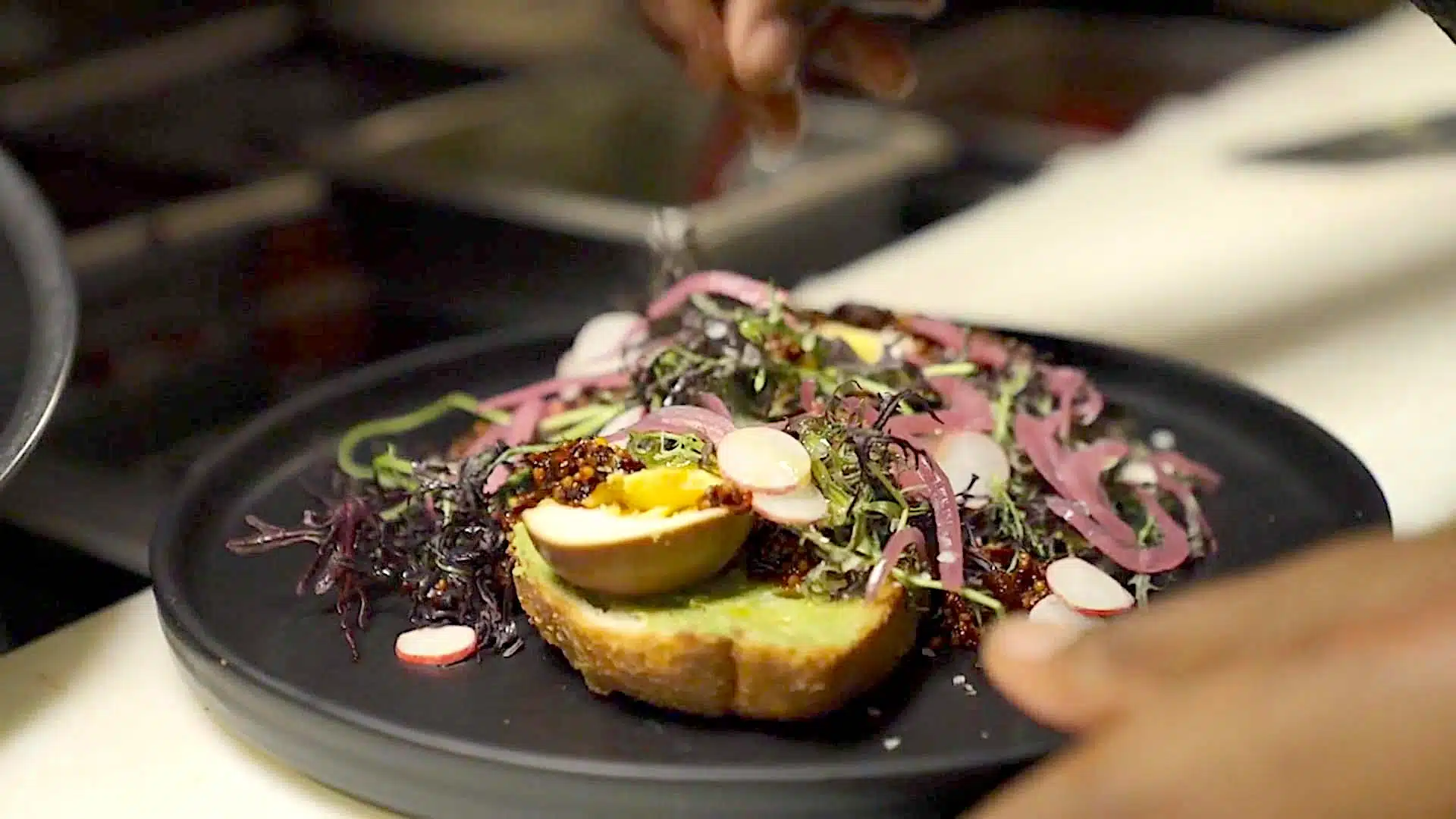 A plate with a slice of bread topped with a boiled egg, purple seaweed, radishes, pickled red onions, and greens, with a hand sprinkling seasoning in the background.