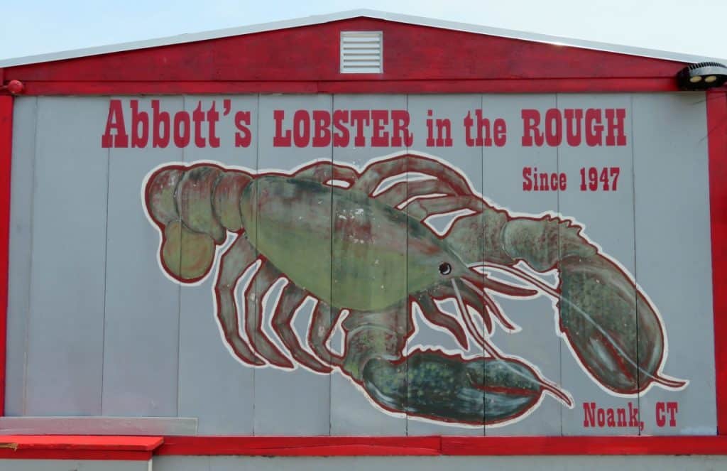 Signage for "Abbott's Lobster in the Rough" featuring a large painted lobster and text indicating it has been in business since 1947, located in Noank, Connecticut.