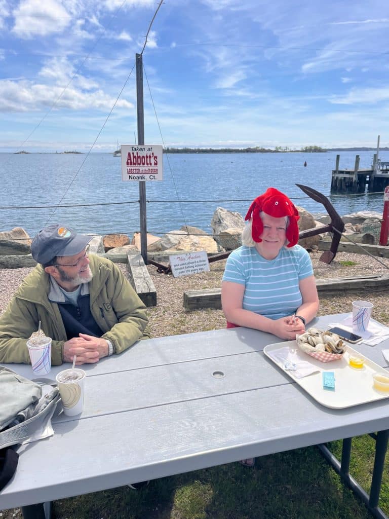 Two people sitting at a picnic table near the water, one wearing a red lobster hat. There are food trays on the table, and a sign in the background reads "Welcome to Abbott's, Noank, CT.