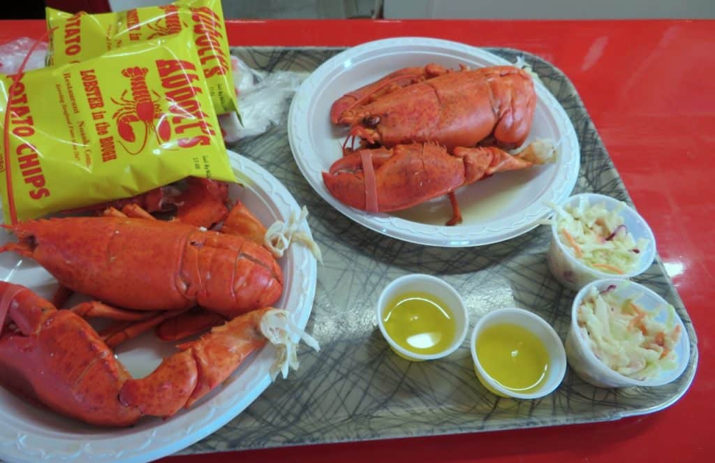 Two plates of whole cooked lobsters with two bowls of melted butter, three small bowls of coleslaw, and two bags of potato chips on a tray.