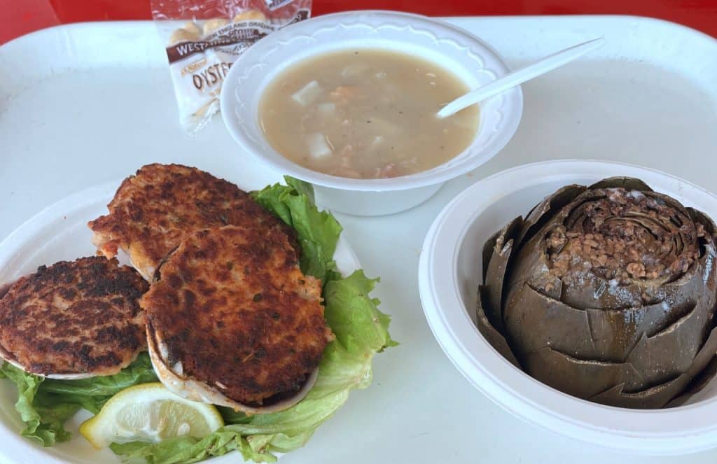 A tray with a bowl of clam chowder, three crab cakes on lettuce with a lemon wedge, a whole steamed artichoke, and a pack of oyster crackers.