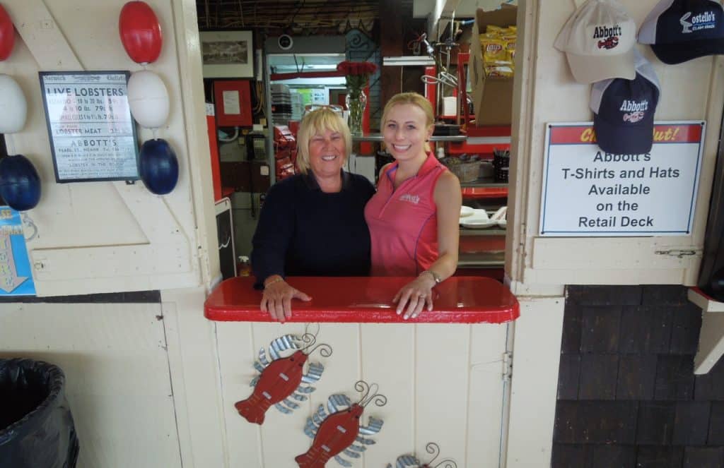 Two women stand behind a counter at a retail stall. Signs advertise live lobsters and merchandise for sale. Neatly arranged hanging items are visible in the background.