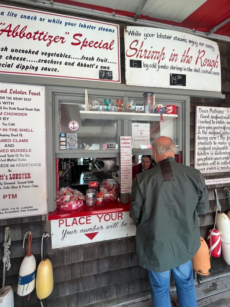 A man in a green jacket orders food from a window at a seafood stand. The menu on the left lists various seafood dishes like lobster, chowder, and clams.