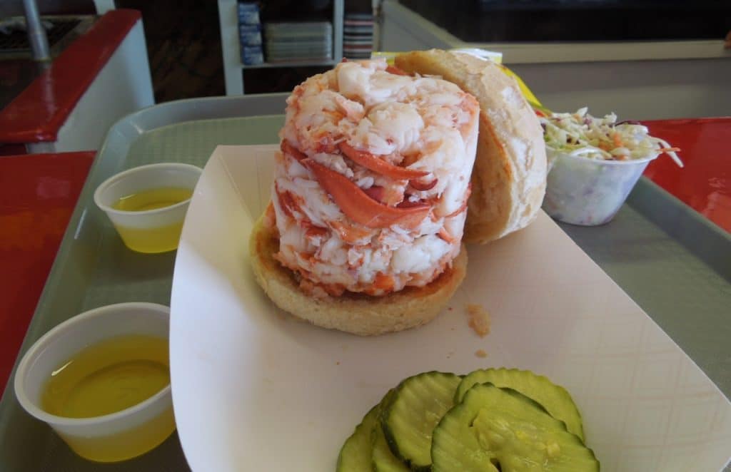 A sandwich with a large portion of lobster meat on a bun, served on a tray with pickles and small cups of melted butter, with coleslaw in the background.