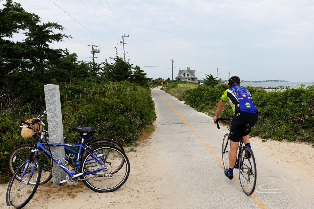 A cyclist rides on a paved bike path beside the beach, with another bicycle parked on the sand nearby. Trees and bushes line the path, and a building is visible in the distance.