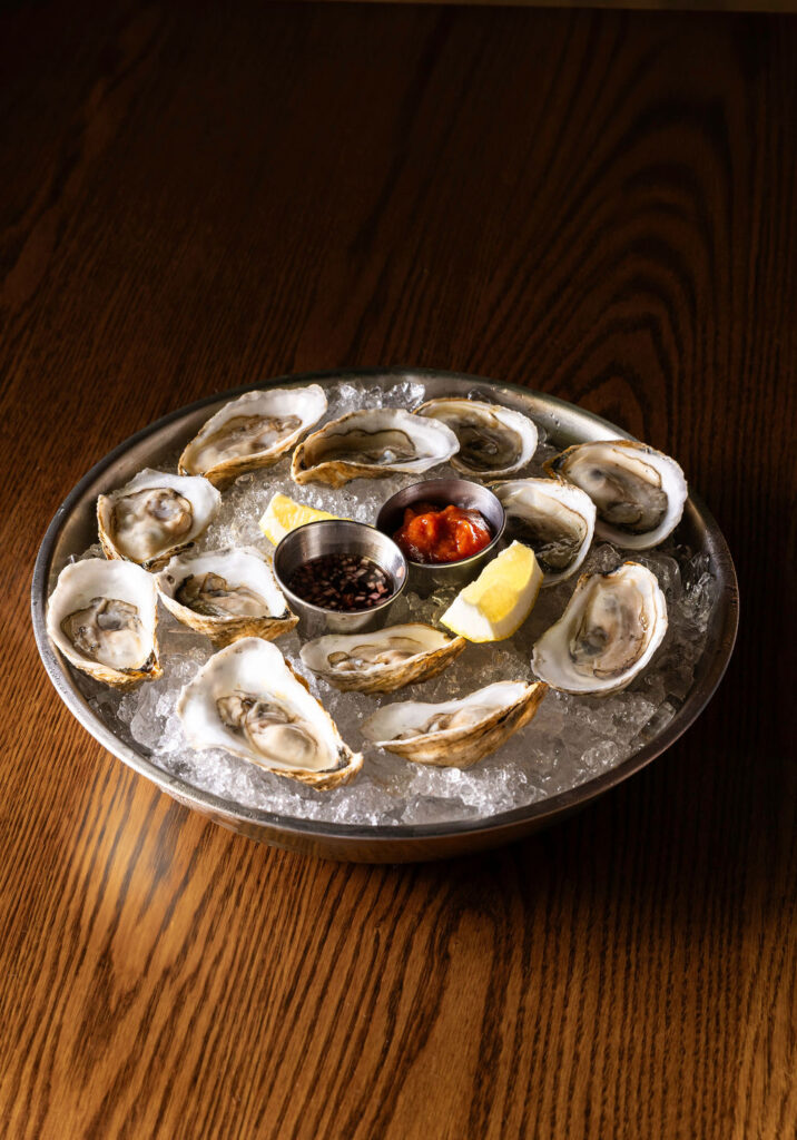 At Dry Dock in Portland, Maine, a metal plate of raw oysters on ice, served with lemon wedges, cocktail sauce, and a small container of mignonette on a wooden table.