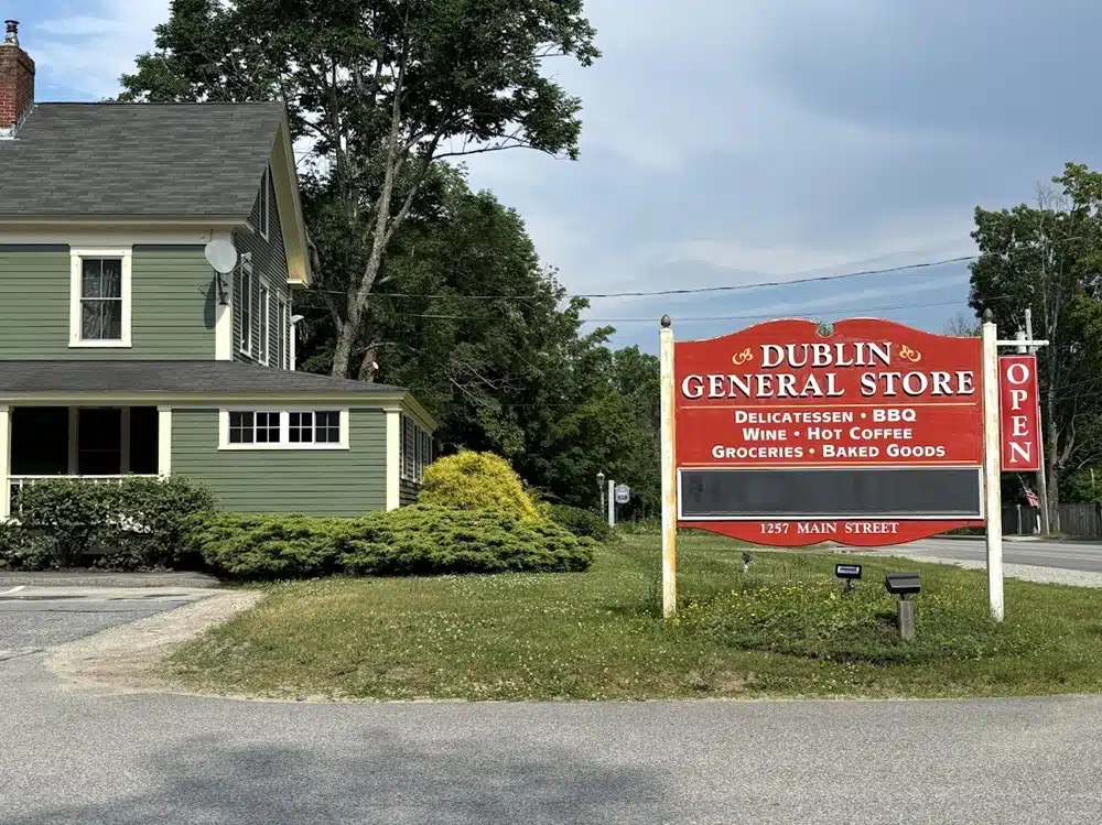 A green building houses the Dublin General Store with a red sign. The sign lists items like delicatessen and groceries. An "OPEN" sign hangs beside it. Address: 1257 Main Street. Trees in the background.