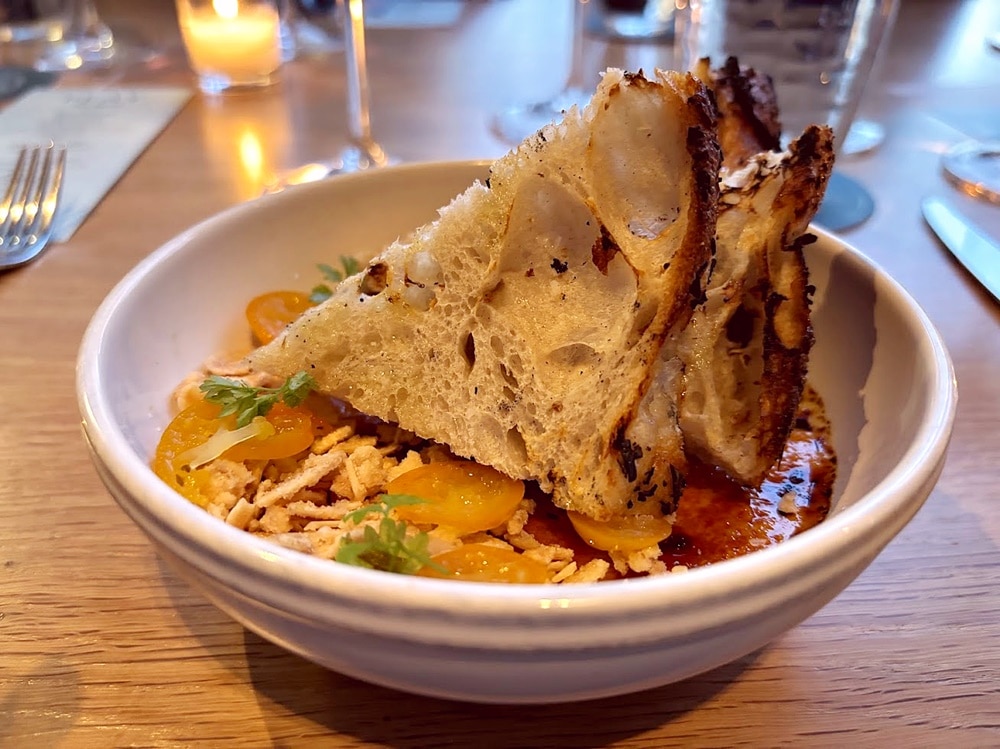 A bowl of food with cherry tomatoes, shredded meat, and slices of toasted bread served on a wooden table with glasses and a candle in the background.