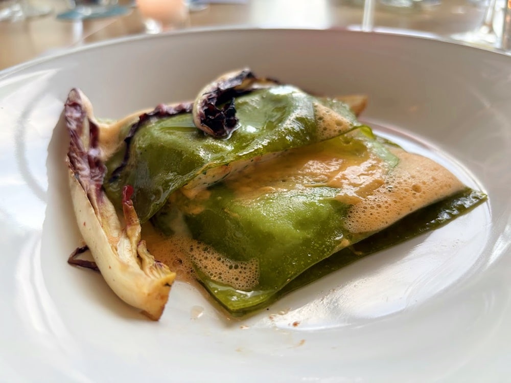 A dish of green ravioli topped with a frothy sauce, accompanied by wilted leafy greens, served on a white plate.