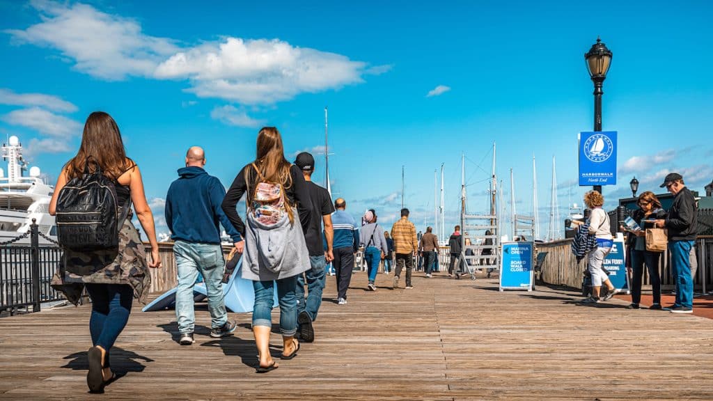 People walking along a wooden pier on a sunny day with blue skies, various signs visible, and boats docked in the background.