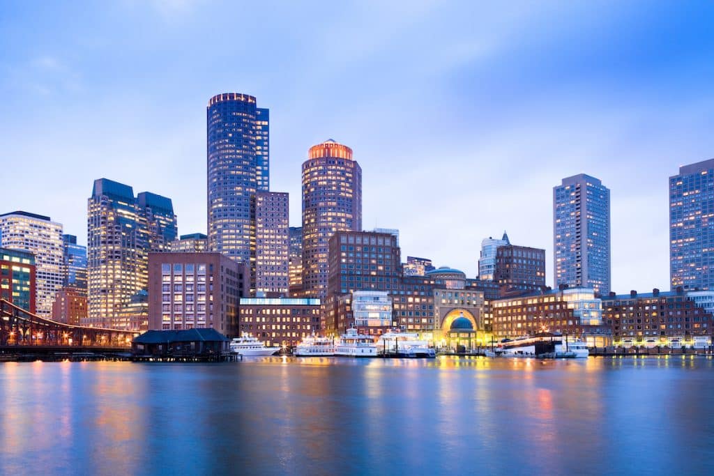 A city skyline at dusk with tall buildings illuminated by lights, reflected in a body of water in the foreground.