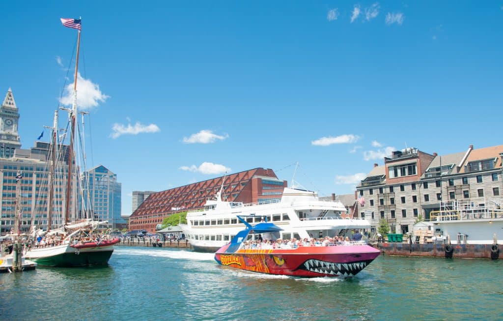 Colorful speedboat and classic sailboat in an urban harbor with modern buildings in the background under a clear blue sky.