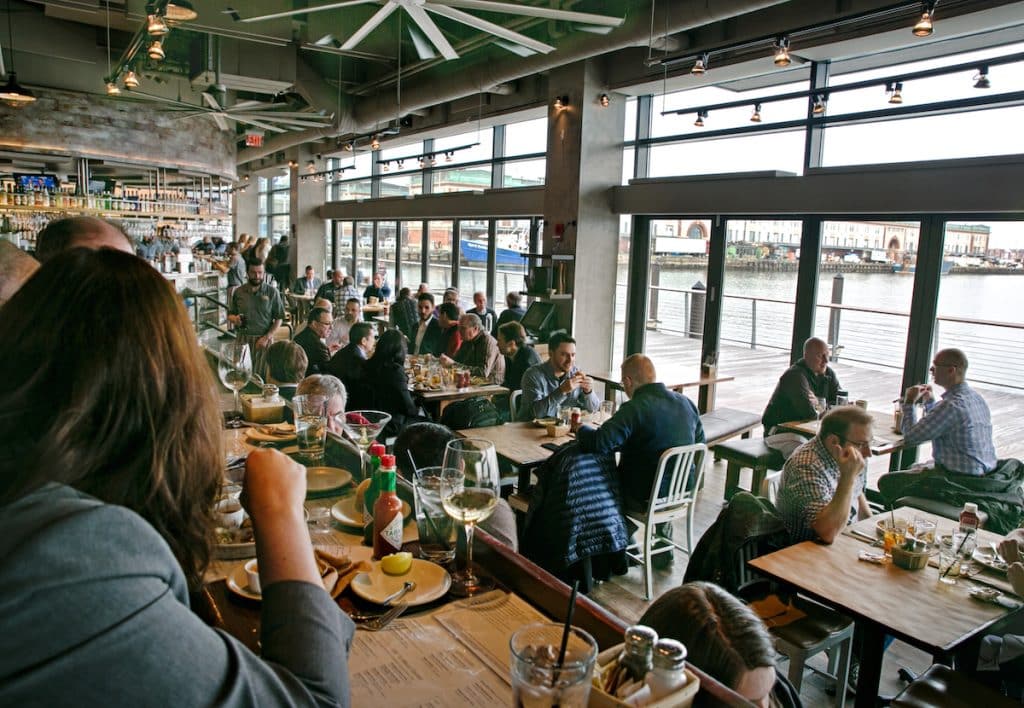 A bustling restaurant with people dining at tables and a bar, featuring large windows offering a view of a waterfront outside.