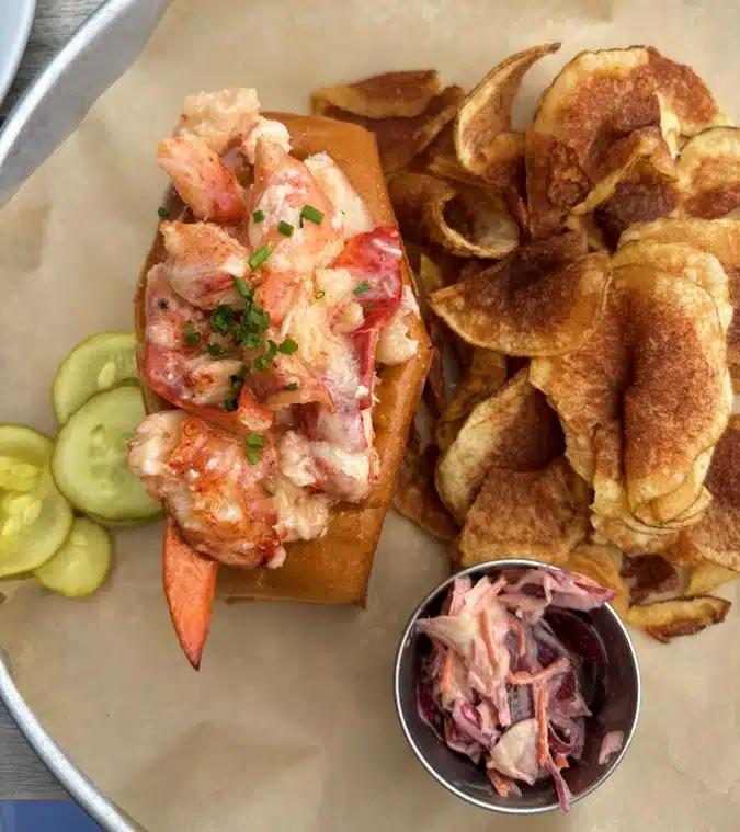 A lobster roll garnished with chives, accompanied by a side of potato chips, pickles, and coleslaw in a small metal dish, all served on a parchment-lined tray.