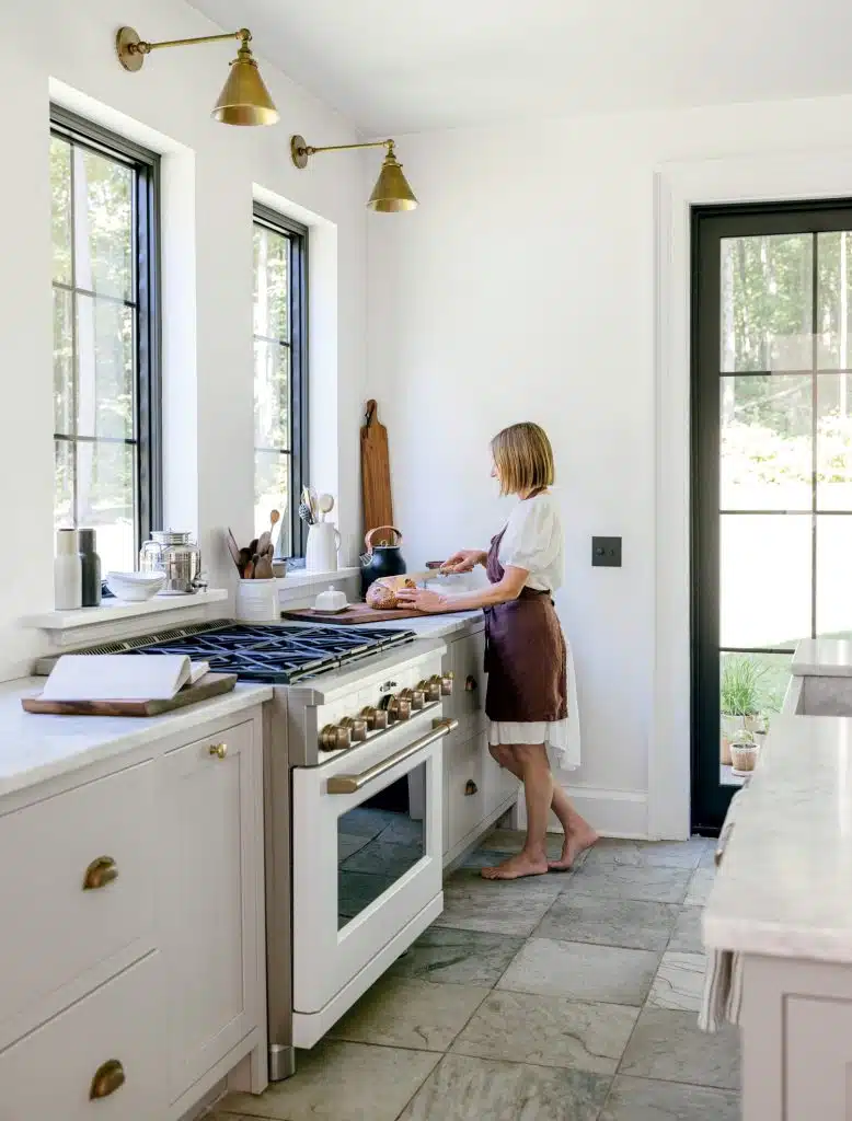 A person stands cooking at a kitchen counter, surrounded by white cabinetry, with large windows and a door letting in natural light.