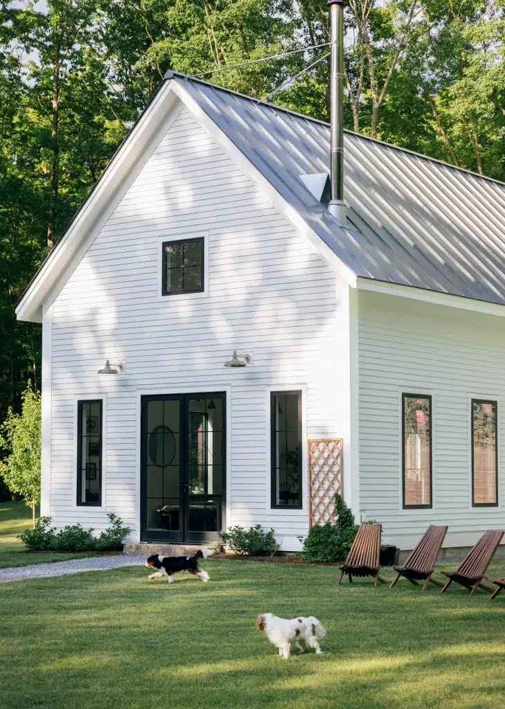 A white, modern farmhouse with a metal roof and black-framed windows sits amidst a green lawn surrounded by trees. Two dogs are playing on the grass near a line of wooden chairs.