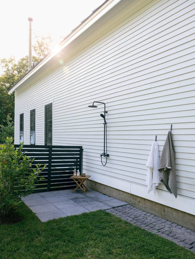 Outdoor shower with black fixtures on the side of a white house, with towels hanging on hooks and a small wooden table with toiletries nearby.