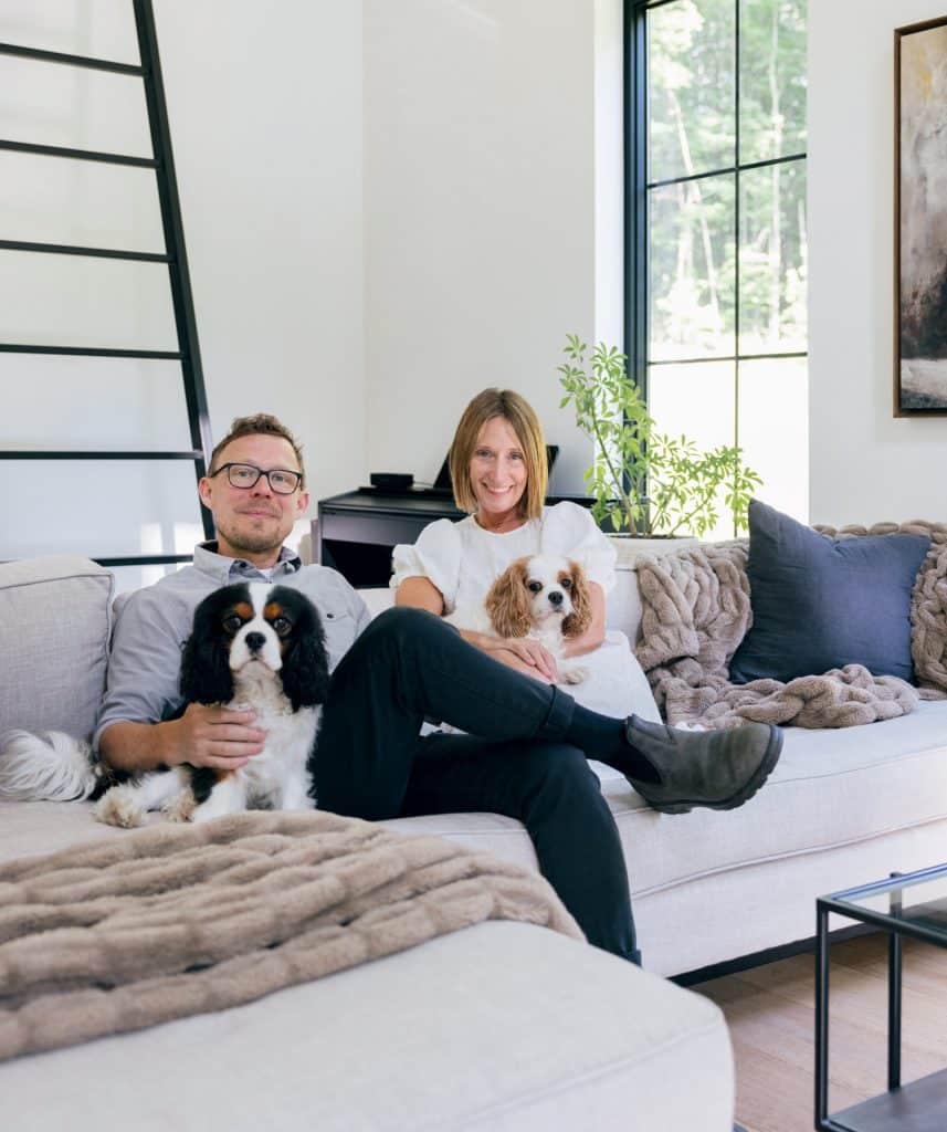 A man and a woman are sitting on a sofa in a well-lit living room, each holding a dog. The room features large windows, a ladder, and cozy furnishings.