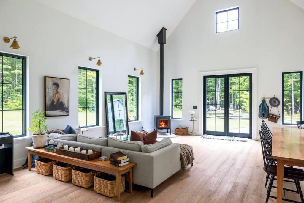 Spacious living room with large windows, a fireplace, a gray sectional sofa, and wooden accents. The room has tall ceilings and natural light, featuring a mix of modern and rustic decor elements.