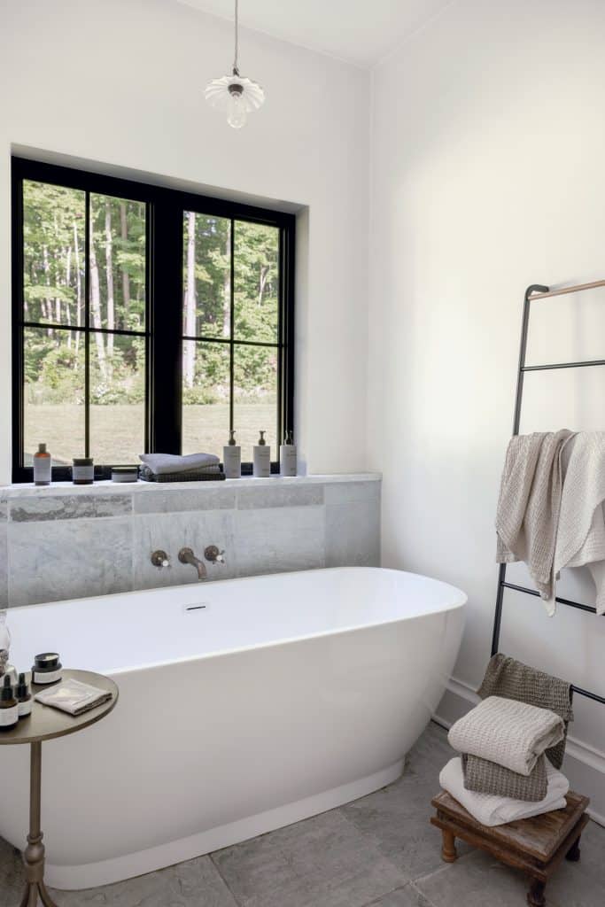 Modern bathroom with a white bathtub, a small round table with toiletries, towels on a ladder shelf, and a large window providing natural light.