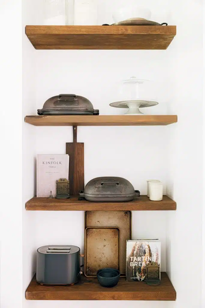 Open shelving with wooden boards displaying various kitchen items, including cast iron pots, a toaster, a cake stand, cooking books, and small bowls against a white wall backdrop.