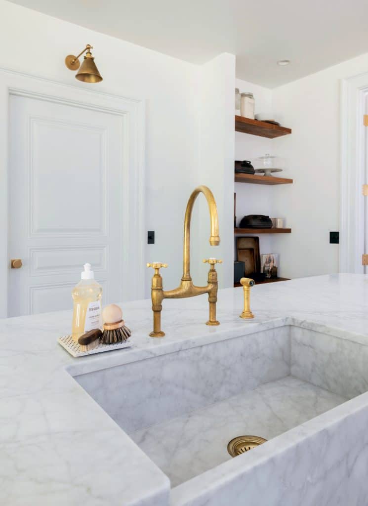 A marble kitchen island with a brass faucet, soap dispenser, brushes, and shelves with various items is shown. There is a white door and wall sconce in the background.