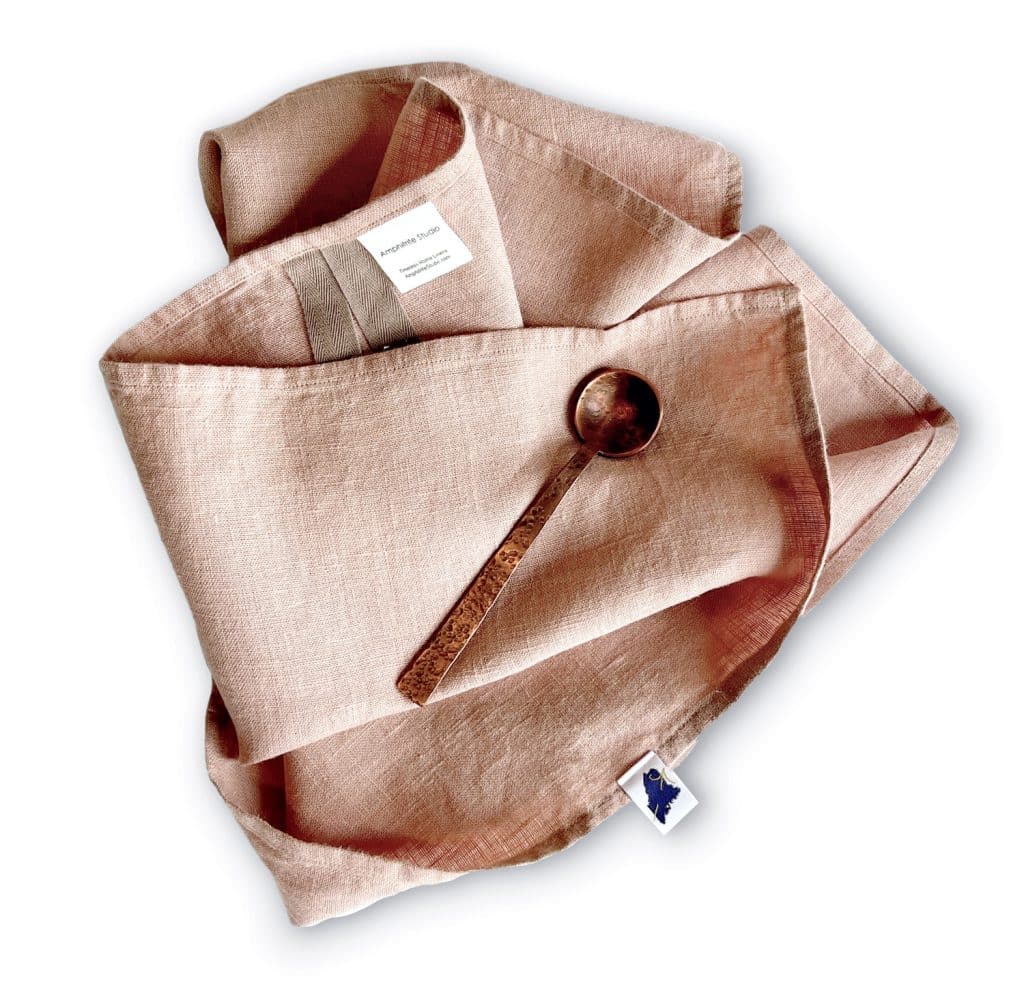 A folded beige linen cloth with a label and pocket, containing a tarnished metal spoon rests on a white background.