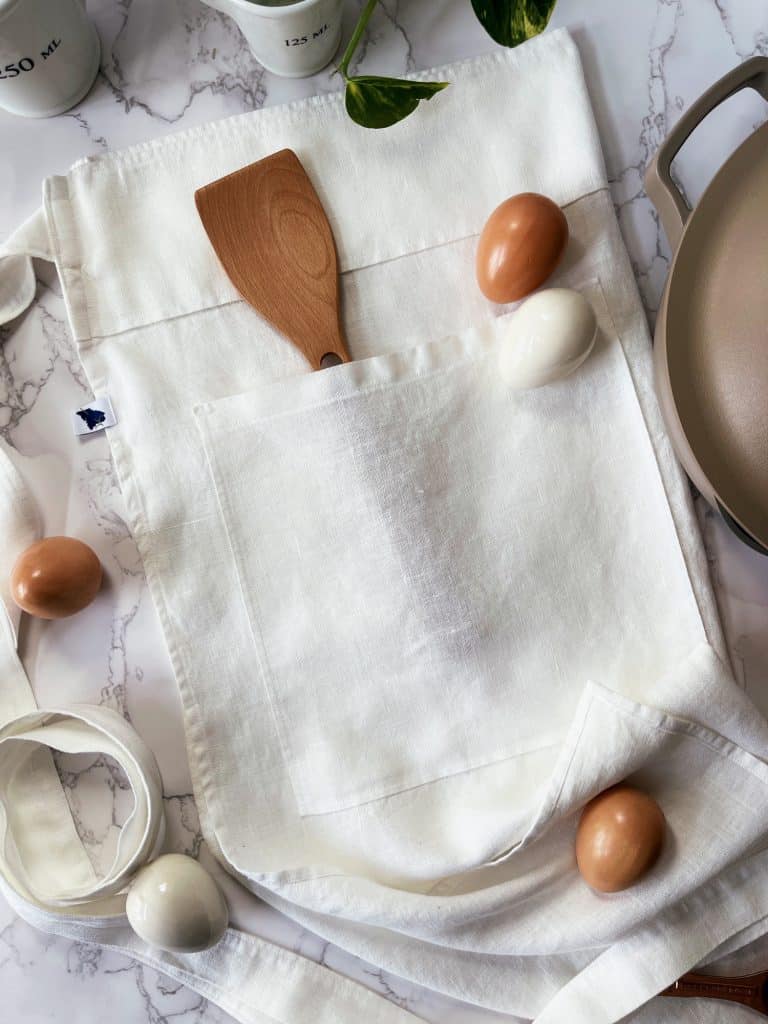 A white apron with a wooden spatula in its front pocket lays on a marble surface. Five eggs, four brown and one white, are scattered around the apron. A pan is partially visible to the right.