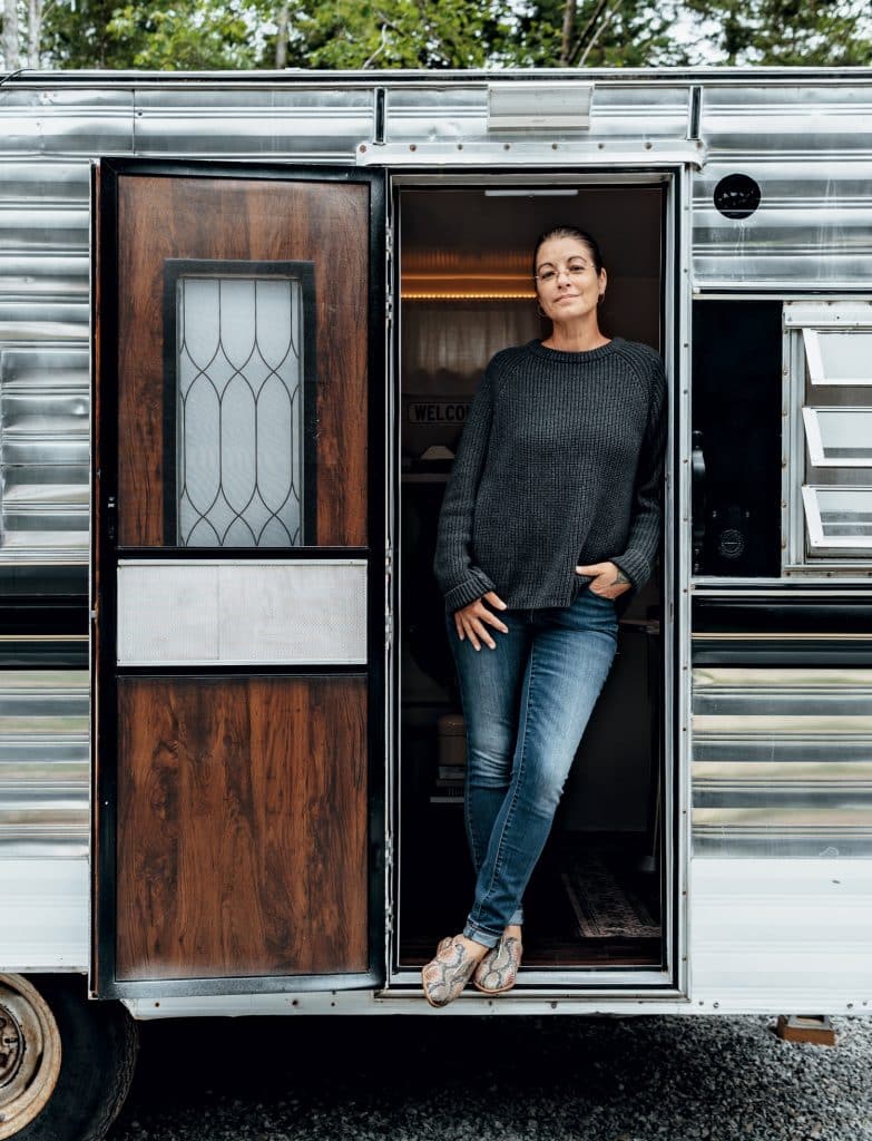 A person stands at the entrance of a vintage-style trailer, wearing a dark sweater, jeans, and sandals, with one hand on the door frame and the other in their pocket.