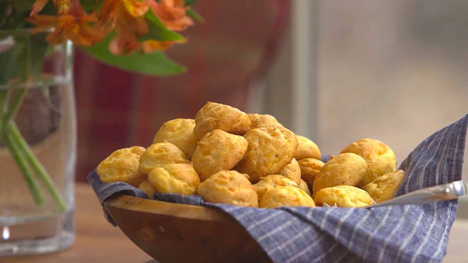 A wooden bowl lined with a blue cloth holds several small, round cheese bread rolls. A glass vase with orange flowers is visible in the background.