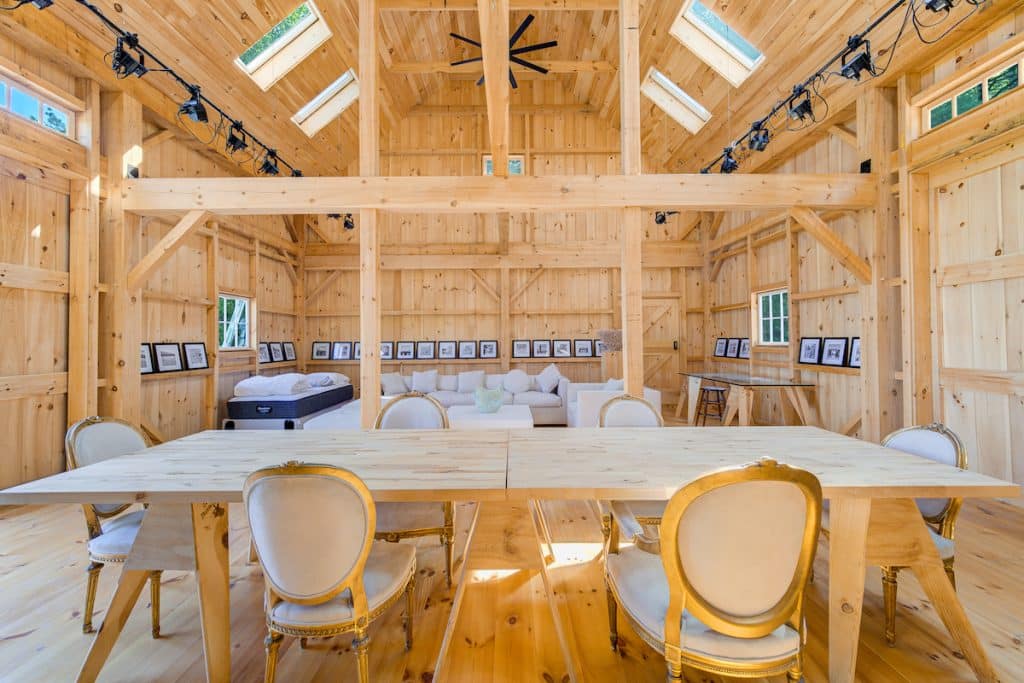 A spacious wooden interior with a large table and chairs in the foreground, a sofa and framed photos on shelves against the back wall, and ceiling skylights.