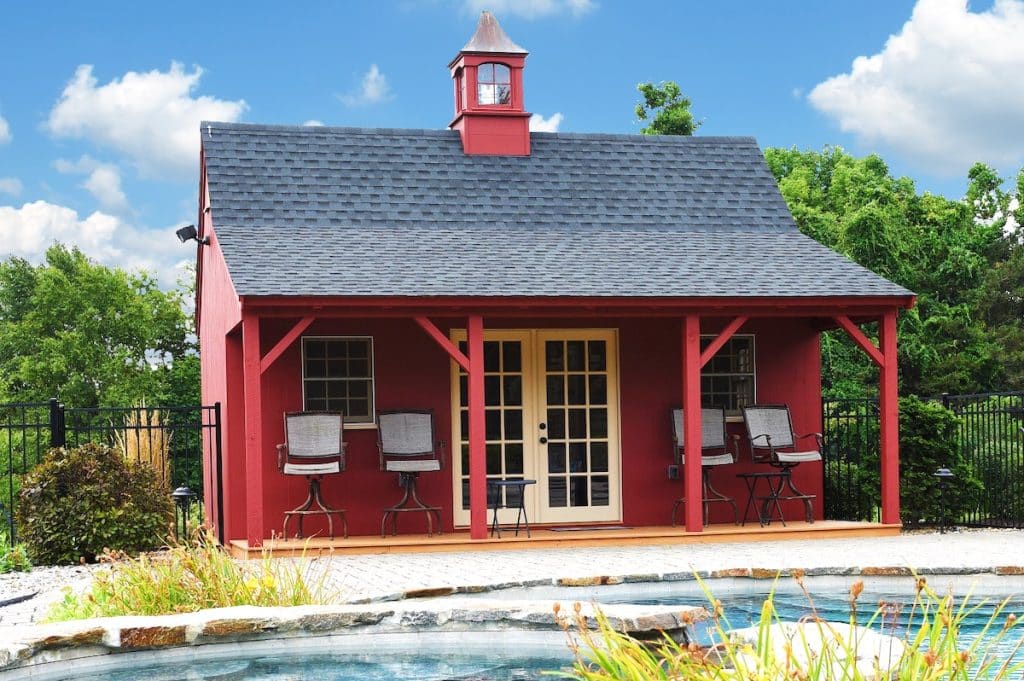 A small red building with a porch, black roof, and cupola stands in front of a fenced-in area with lush greenery and a pond in the foreground. Four chairs are arranged on the porch.