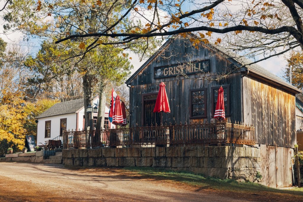 A rustic wooden building with an outdoor patio featuring red-striped umbrellas. Trees with autumn leaves surround the structure. Dirt path and another small building are visible.