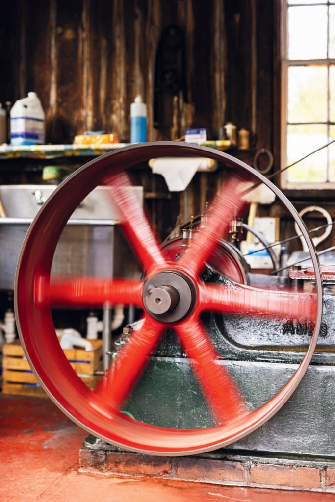 A red flywheel spins rapidly on an old, green industrial machine in a workshop with wooden walls and various bottles and tools on shelves in the background.