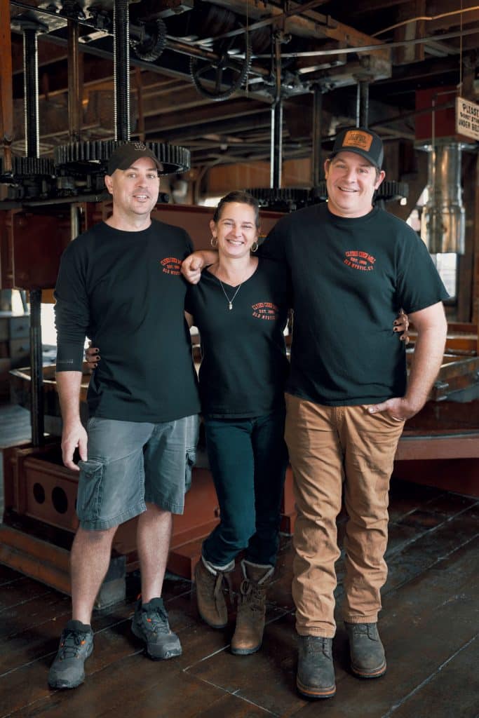 Three people wearing matching black shirts and hats stand arm-in-arm in what appears to be an industrial or warehouse setting.