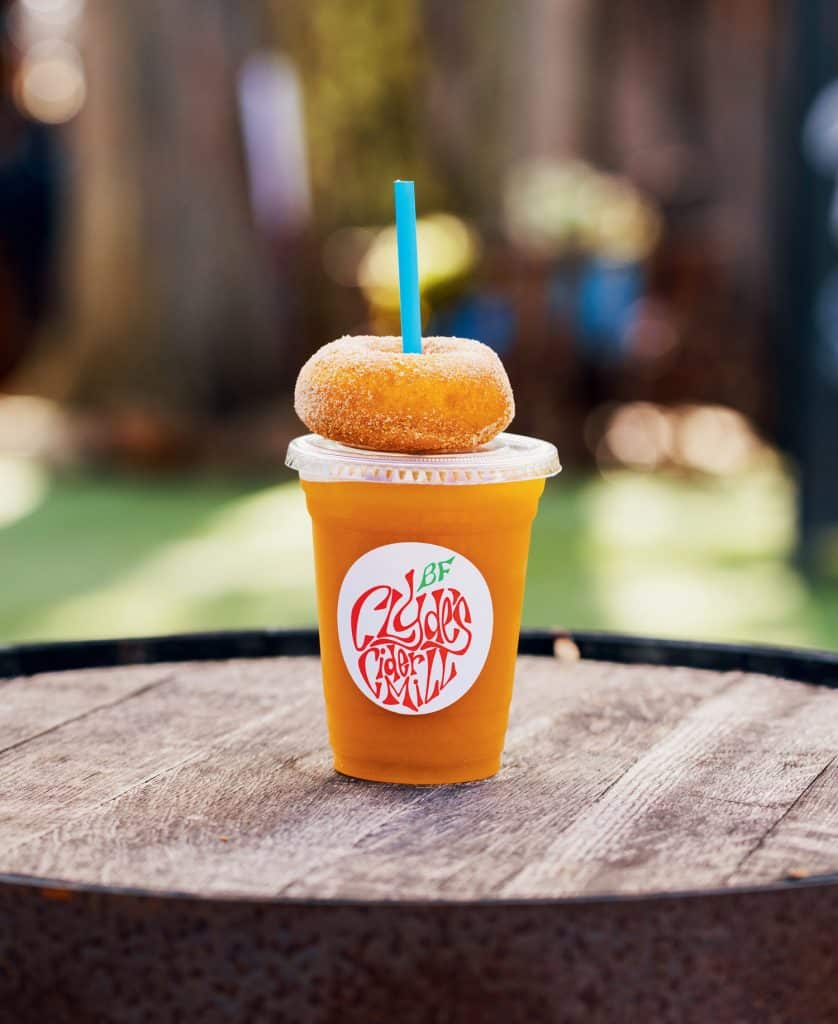 A plastic cup filled with an orange beverage and topped with a sugar-coated donut. A blue straw is inserted through both the donut and the cup lid. The cup has a logo that reads "Clyde's Cider Mill.