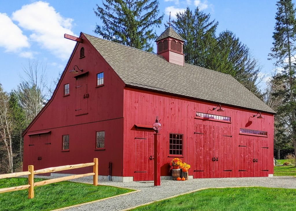 A red barn with a gabled roof stands against a backdrop of trees. The barn has multiple windows and doors, and a wooden fence runs alongside it. A potted plant with orange flowers is near the entrance.