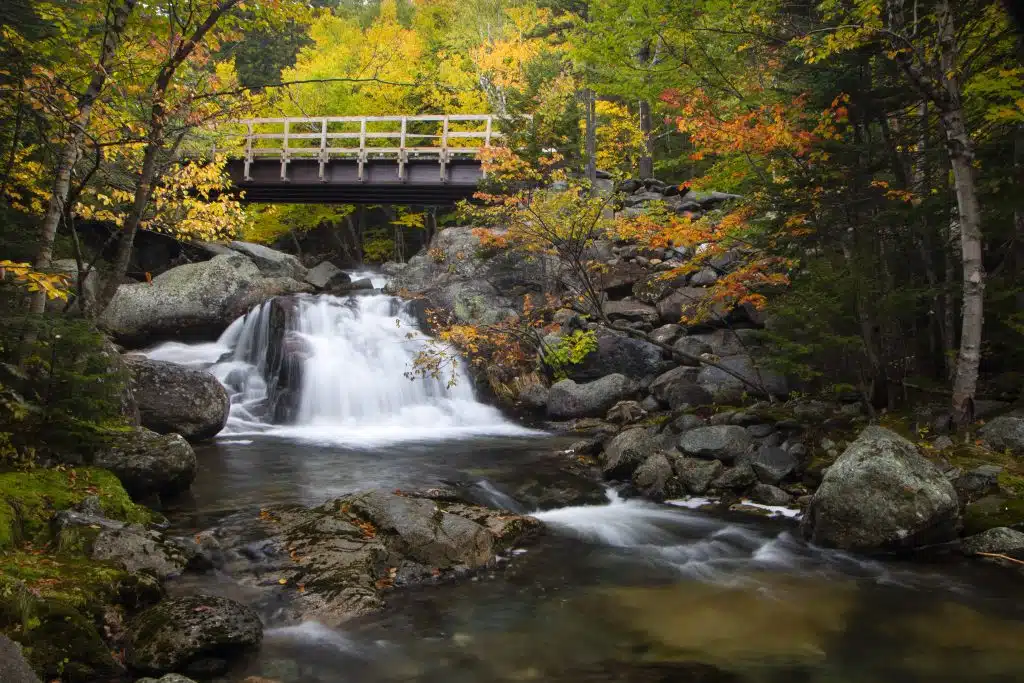 A small waterfall flowing under a wooden bridge surrounded by rocks and trees with autumn foliage.